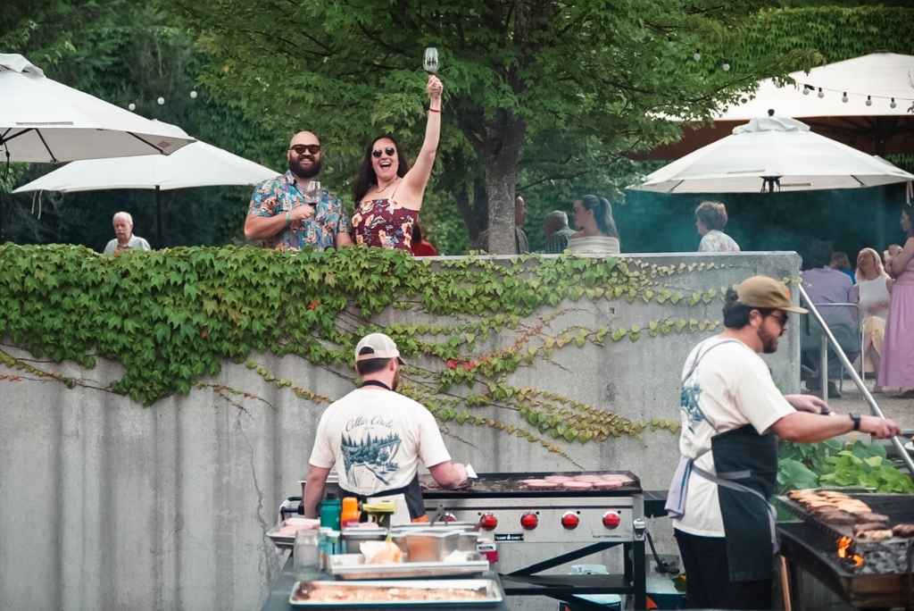 Guests toast with glasses of wine at a summer wine club event at Novelty Hill-Januik while chefs cook outdoors on the barbecue.